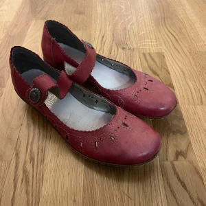 Röda ballerinaskor från Rieker - Snygga röda ballerinaskor från Rieker i skinn med dekorativa broderier och rem med knappdetalj. Rund tå och låg klack ger en klassisk look. Perfekta för dig som vill ha något unikt och bekvämt till vardags. Säljer pga att de är för stora! De är jättefina och passar till nästan allt plus att det är äkta skinn .                                                                         OBS! Köparen står för frakt!