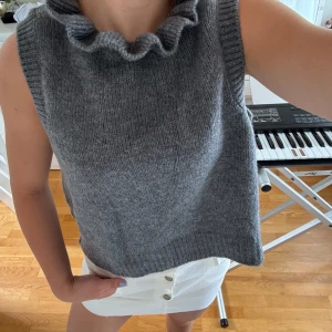 Grå stickad väst med volangkrage från YAS - Säljer en grå stickad väst från VAS med en snygg volangkrage. Västen är ärmlös och har en rak, lite loose passform. Perfekt att styla över en skjorta eller t-shirt för en trendig lager-på-lager-look.