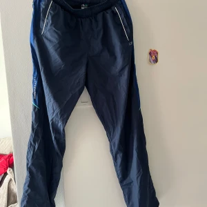 Mörkblå täckbyxor från LS - Mörkblå täckbyxor från LS med elastisk midja och vita detaljer vid fickorna. Byxorna har blå sidopaneler och en tunn turkos linje baktill. Perfekta för kalla dagar eller utomhusaktiviteter.