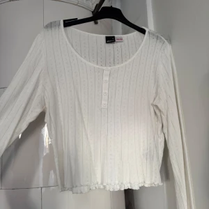 Vit ribbad topp från Gina Tricot - Supersöt vit långärmad topp från Gina Tricot med ribbat mönster och små håldetaljer. Rund halsringning, fyra knappar framtill och vågig kant nertill. 