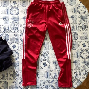 Röda Ajax träningsbyxor Adidas - Snygga röda träningsbyxor från Adidas med Ajax Amsterdam-logga och tre vita ränder längs sidorna. Byxorna har elastisk midja och smal passform, perfekta för fotboll eller träning. Materialet är mjukt och lätt polyester. 