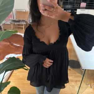 Svart blus med volang och vida ärmar - Supersöt unik svart blus med djup v-ringning och volangkant. Blusen har vida, plisserade långärmar och en flowy passform. Tillverkad i ett tunt, lätt material som faller snyggt. 