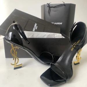 Svarta Saint Laurent sandaler med YSL-klack - Superlyxiga svarta sandaler från Saint Laurent i lackat skinn med ikonisk guldig YSL-klack. Snygg fyrkantig tå, smal rem över tårna och justerbar rem runt ankeln. Perfekta för dig som vill sticka ut med en statement-klack och elegant design.
