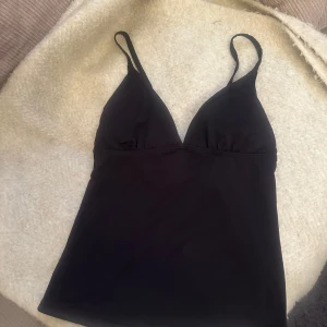 Bikini  - En svart linnetopp med smala, justerbara axelband och djup v-ringning. Toppen har en enkel och stilren design med lätt figurnära passform. Perfekt att matcha med jeans eller kjol för en snygg look.