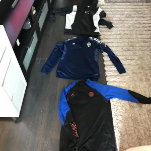 Nike Air hoodie, Adidas Sverige och PSG-tröja - Tre snygga tröjor: svart och vit Nike Air hoodie med huva, blå långärmad Adidas Sverige-tröja med broderad logga och axelränder, samt svart och blå PSG-tröja från Jordan med röd logga och half zip. Perfekta för fotboll eller chill. Skicka dm för mer info om tröjorna 1st för  200-300 pris diskuteras
