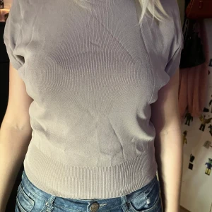 Beige ribbad topp med kort ärm - En stilren beige topp med korta ärmar och ribbad nederkant. Toppen har en enkel, rak passform och är tillverkad i ett mjukt material som känns skönt mot huden. Perfekt att matcha med jeans eller kjol för en clean look. Finns ingen lapp tyvärr för den är såld second hand 