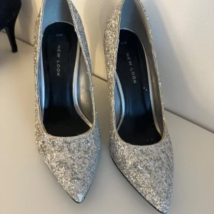Glittriga silverpumps från New Look - Snygga pumps från New Look i silver med glittrig yta och spetsig tå. Perfekta för dig som vill ha extra bling på fötterna. Insidan är fodrad i silverfärgat material. Köpta på Sellpy men själv aldrig använt. Dem är lite slitna på kanterna och lite på ena tån (se bild).