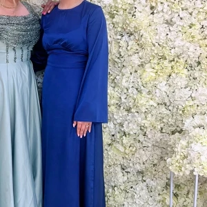 Marinblå långklänning med vida ärmar - Säljer en elegant marinblå långklänning med rund halsringning och långa, vida ärmar. Klänningen har en markerad midja och är tillverkad i ett slätt, glansigt tyg som ger en lyxig känsla. Perfekt för dig som vill sticka ut med stil. Snörning finns där bak. 