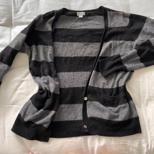 Randig kofta - Kofta i svart och grått med breda ränder. Modellen har långa ärmar, två fickor framtill och stängs med två guldiga knappar. 