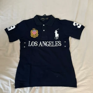Mörkblå Polo Ralph Lauren t-shirt M - Mörkblå t-shirt från Polo Ralph Lauren med klassisk pikékrage, broderad logga och Los Angeles-tryck framtill. Stora vita siffror på ärm och rygg, samt broderad emblem på bröstet. Tillverkad i bomull och har en normal passform.