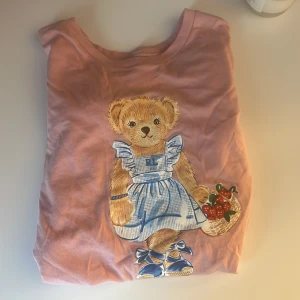 Rosa Polo Bear t-shirt från Ralph Lauren - Supersöt rosa t-shirt från Ralph Lauren med Polo Bear-tryck framtill. Björnen har blå klänning och håller en korg med jordgubbar. Klassisk passform och korta ärmar, perfekt för dig som gillar gulliga detaljer och ikoniska prints.