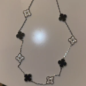 Silverfärgat halsband med klöverhängen - Stilrent halsband i silverfärg med växelvis svarta och glittriga fyrklöverformade hängen. Kedjan är tunn och elegant, och klöverhänget har en fin pärlad kant. Perfekt accessoar för att lyfta din outfit med en touch av lyx.