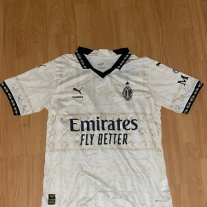 Fitbollströja - Fotbollströja i vitt med subtilt mönster och svarta detaljer. Puma-logga, klubbmärke och sponsortryck på bröstet. Svart krage och ärmslut med mönstrad kant. Perfekt för dig som vill visa ditt stöd för Milan.