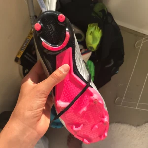 Nike Mercurial fotbollsskor silver/rosa - Snygga Nike Mercurial fotbollsskor i silver med svart och rosa detaljer. Skorna har svart snörning, högre strumpliknande skaft och rosa dobbar för grymt grepp på planen. Lätta och smidiga för snabba rörelser, perfekt för dig som vill sticka ut på matchen. 