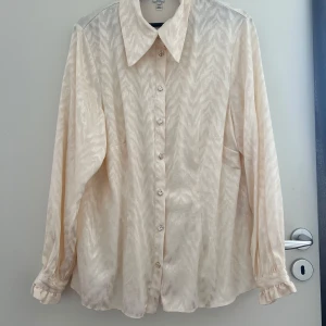 Beige skjorta från River Island, stl 44 - Snygg cremevit skjorta från River Island med subtilt zickzackmönster. Skjortan har klassisk krage, knappar framtill och långa ärmar med volangdetalj vid ärmslut. Tillverkad i en mjuk blandning av viskos och polyester som ger ett lyxigt skimmer.
