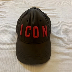 Svart keps Dsquared2 ICON rosa text - Svart keps från Dsquared2 med böjd skärm och justerbart spänne bak. Framtill står det 'ICON' i stora röda broderade bokstäver och baktill 'DSQUARED2' i samma stil. Kepsen är i bomull och har klassisk sexpanelsdesign. Kepsen är aningen solblekt