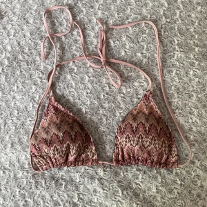 Mönstrad bikiniöverdel med snäckberlock - Supersnygg bikiniöverdel i rosa, beige och bruna toner med ett unikt mönster och glittriga detaljer. Triangelformad modell med knyt i nacken och ryggen. Söt snäckformad berlock framtill. Tillverkad i 100% polyester. Använd några gånger, storlek M. 