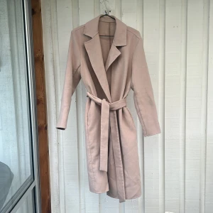 Beige kappa med bälte - Snygg beige kappa med klassisk krage och bälte i midjan. Lång modell med raka ärmar och stilren design, perfekt för dig som gillar en clean och trendig look