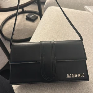 Svart axelväska från Jacquemus - Säljer en stilren svart axelväska från Jacquemus i slätt skinn. Väskan har en rektangulär form, minimalistisk design och ett diskret Jacquemus-logo i metall på framsidan. Invändigt finns ett fack för kort eller småsaker. Perfekt för dig som gillar clean och lyxig vibe.