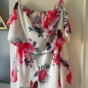Vit blommig offshoulder sommarklänning - Superfin vit kortklänning med stora rosa och röda blommor, offshoulder-modell och flowig passform. Perfekt för varma dagar, med romantiska volanger och tropiska blad i mönstret. Lätt och luftig, passar dig som vill sticka ut med färg och mönster. Den har shorts inuti som inte syns, så man behöver inte vara rädd för att den flyger upp!
