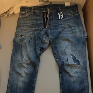Dsquared2 blå slitna jeans med dragkedja - Blå jeans från Dsquared2 med coola slitningar och hål på benen, samt en stor silverfärgad dragkedja framtill. Jeansen har en tvättad look och klassiska bakfickor. Perfekt för dig som gillar streetstyle och vill sticka ut.