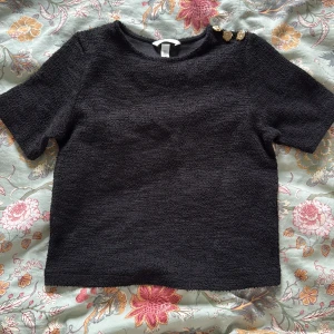 Svart topp med guldknappar H&M - Svart kortärmad topp från H&M med strukturerad yta och tre dekorativa guldknappar på ena axeln. Enkel och stilren modell med rund halsringning, perfekt för att lyfta en outfit med lite extra detaljer.