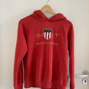 Roströd hoodie från GANT med tryck - Roströd hoodie från GANT med broderat logotyp-tryck och sköld på bröstet. Klassisk känguruficka framtill, huva och ribbade muddar. Mjuk bomullsblandning och långärmad modell, perfekt för en chill och sportig look.