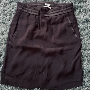 Svart kortkjol från Filippa K  - Svart kortkjol från Filippa K i storlek XS. Kjolen har elastisk midja med snörning och två fickor framtill. Enkel och stilren design som passar perfekt till sneakers eller boots. Materialet känns mjukt och lätt, perfekt för en chill och snygg look. Skriv om du undrar något🩷