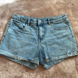 Ljusblå jeansshorts från Kappahl - Snygga ljusblå jeansshorts från Kappahl med klassisk femficksdesign och råa, fransiga benslut. Perfekta för sommaren med sin avslappnade vibe och bekväma passform. Gjorda i mjukt denimtyg och har bälteshällor och knappstängning framtill.