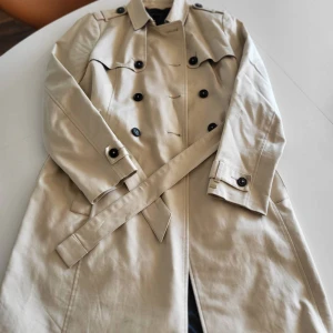 Beige trenchcoat från Tommy Hilfiger - Stilren beige trenchcoat från Tommy Hilfiger med klassisk dubbelknäppning, bälte i midjan och snygga detaljer vid ärmslut. Jackan har långa ärmar och är tillverkad i bomull, perfekt för vår och höst. En tidlös favorit som lyfter varje outfit.