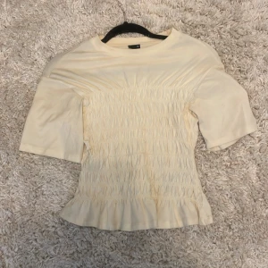 Gul smockad topp från Gina Tricot - Supersöt gul topp från Gina Tricot i smockad modell med korta ärmar och volangkant nertill. Toppen har rund halsringning och är gjord i mjukt material som sitter bekvämt. Perfekt för dig som gillar en feminin och trendig stil.