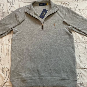 Grå half zip tröja från Polo Ralph Lauren - Snygg grå långärmad tröja från Polo Ralph Lauren med klassisk half zip-dragkedja och broderad logga på bröstet. Tillverkad i mjuk bomull, perfekt för lager-på-lager och chill stil. Passformen är normal och tröjan har ribbade muddar.