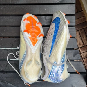 Nike mercurial vapor - Säljer ett par Nike fotbollsskor med en iögonfallande blå swoosh och orange detaljer på sulan. Skorna har snörning och är designade för optimal prestanda på planen.