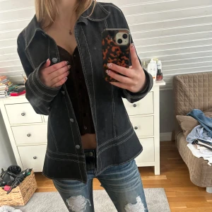 Svart jeansjacka med kontrastsömmar - Snygg svart jeansjacka med vita kontrastsömmar. Jackan har en klassisk krage och knappar framtill. Perfekt till vår och sommar mm. S/M