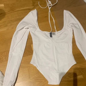 Vit långärmad bodysuit från H&M - Säljer en stilren vit bodysuit från H&M med långa ärmar och djup v-ringning. Den har en knytning framtill som en extra detalj. Perfekt för att styla med jeans eller kjol en kväll ute. Använd en gång. 