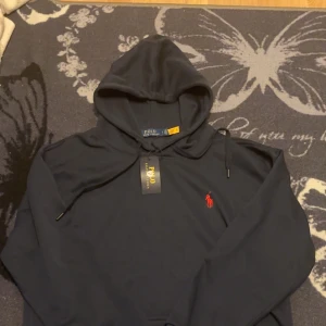Svart hoodie från Ralph Lauren - Säljer en stilren svart hoodie från Ralph Lauren med det klassiska röda logotypen på bröstet. Tröjan har en justerbar huva och långa ärmar med ribbade muddar. Perfekt för en avslappnad stil.