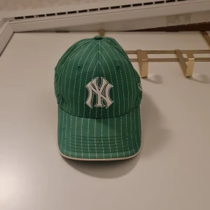 Grön randig keps från New Era - Snygg grön keps med vita ränder och ett broderat emblem framtill. Kepsen är justerbar baktill och har en klassisk design. Perfekt för sportiga tillfällen eller som en stilren accessoar.
