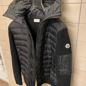Svart cardigan från Moncler - Snygg svart dunväst från Moncler med stickade ärmar och dragkedja framtill. Västen har en quiltad design och en praktisk huva. Perfekt för våren! Mindre i storlekarna då det är så passformen är för dessa jackor.