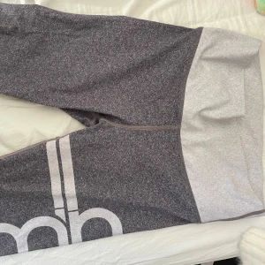 Grå träningsbyxor ifrån AMIN - Snygga grå träningsbyxor med en stor logga på benet. Byxorna är tillverkade i en mjuk och stretchig blandning av polyester och spandex, vilket gör dem perfekta för träning. De har en bekväm passform och elastisk midja.