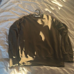 Stone Island tröja - Säljer en snygg mörk grön tröja från Stone Island med deras ikoniska märke på ärmen. Tröjan har långa ärmar och en rund halsringning. Perfekt för en stilren och avslappnad look.