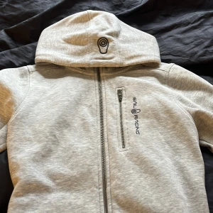 Grå hoodie från Sail Racing - Säljer en stilren grå hoodie från Sail Racing med dragkedja och logotyp på bröstet. Perfekt för en avslappnad stil. Den har en bekväm passform och en praktisk huva.