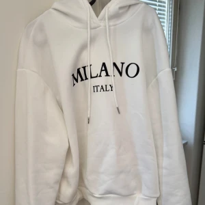 Vit hoodie med texten Milano Italy - Säljer en stilren vit hoodie med texten 'Milano Italy' i svart på bröstet. Den har en klassisk design med huva och dragsko samt ribbade muddar vid ärmslut och nederkant. Perfekt för en avslappnad look. Lite smutsig men något som försvinner i tvätt.