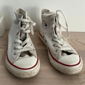 Vita sneakers från Converse - Klassiska vita höga sneakers från Converse med snörning och rund tå. De har den ikoniska röda och blå logotypen på sidan och en röd rand runt sulan. Perfekta för en avslappnad stil.