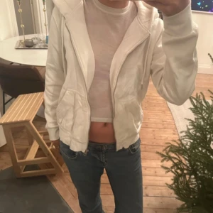Vit hoodie med dragkedja - Säljer en vit hoodie med dragkedja och fickor framtill. Perfekt för en avslappnad stil. Den har långa ärmar och en bekväm passform.