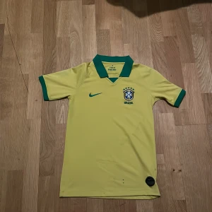 Gul Brasilien fotbollströja från Nike - Säljer en gul Brasilien fotbollströja från Nike med gröna detaljer. Tröjan har korta ärmar och en klassisk krage. Perfekt för fotbollsfans som vill visa sitt stöd för Brasilien! 🇧🇷
