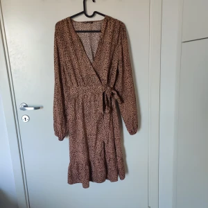 Mönstrad omlottklänning från Shein - Säljer en snygg mönstrad omlottklänning från Shein i beige och svart. Klänningen har långa ärmar och ett knytband i midjan för en smickrande passform. Perfekt för en stilfull look!