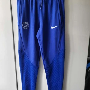  psg byxor - Säljer ett par blå träningsbyxor från Nike med PSG-logga och Dri-FIT-teknologi. Byxorna har en slim fit och är perfekta för sportiga aktiviteter. De har en elastisk midja och är tillverkade i ett syntetmaterial för optimal komfort.