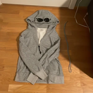 Grå hoodie med glasögon från C.P. Company - Säljer en grå hoodie från C.P.Company Hoodien har används 1 gång. Lite repor på glasögonen 