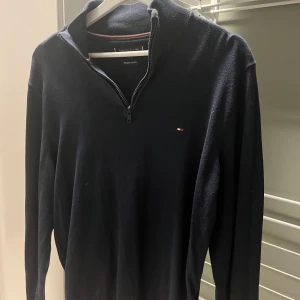 Mörkblå tröja med dragkedja från Tommy Hilfiger - Säljer en stilren mörkblå tröja från Tommy Hilfiger med dragkedja vid halsen. Tröjan har långa ärmar och en liten logotyp på bröstet. Perfekt för en avslappnad och snygg look.