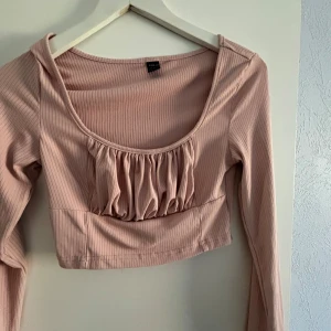 Rosa ribbad topp med volangdetalj - Säljer en söt rosa ribbad topp från Zara med volangdetalj framtill. Toppen är croppad och har långa ärmar, perfekt för en trendig look. Passar bra till både jeans och kjol!